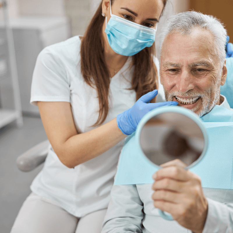 A picture of a senior man smiling looking into a mirror at his dental implants. A dentist sits next to him pointing at his teeth.